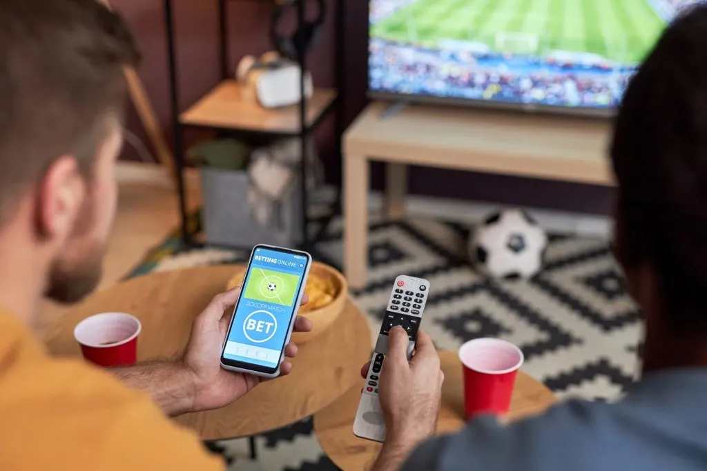 Imagem de dois amigos, de costas, em frente à TV. O da esquerda segura um smartphone aberto em casa de apostas com a mão esquerda e controle remoto com a mão direita. 