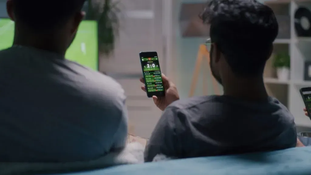Imagem de dois homens assistindo TV, e um deles segurando celular para acompanhar apostas ao vivo
