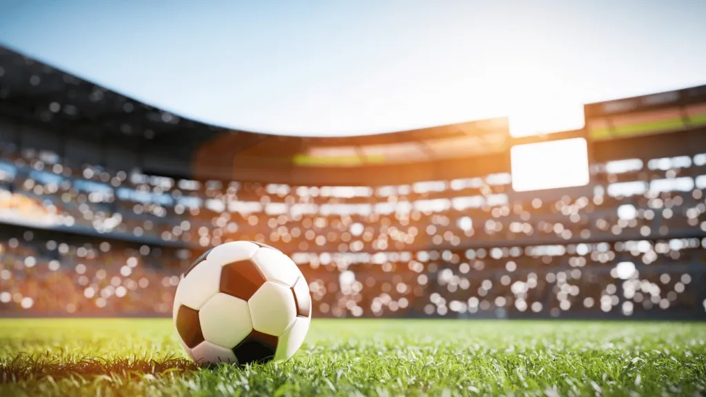 Bola de futebol clássica, em preto e branco, em gramado de estádio de futebol. Ao fundo, pode-se ver holofotes na arquibancada, fora de foco. 