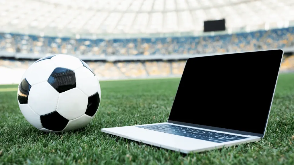Bola de futebol clássica, em branco e preto, ao lado de notebook com tela apagada, ambos na superfície de um gramado de campo de futebol. 