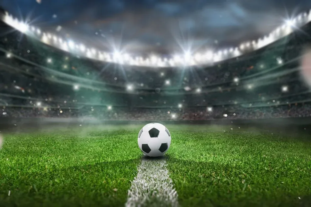 Imagem gráfica de bola de futebol em cima de linha branca que demarca o gramado do campo de futebol. Ao fundo, estádio com arquibancada lotada.