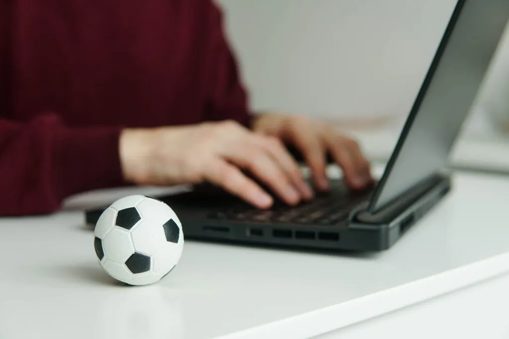 Pessoa usando notebook, apenas mãos e torso à vista. Ao lado, miniatura de bola de futebol clássica, em branco e preto. 