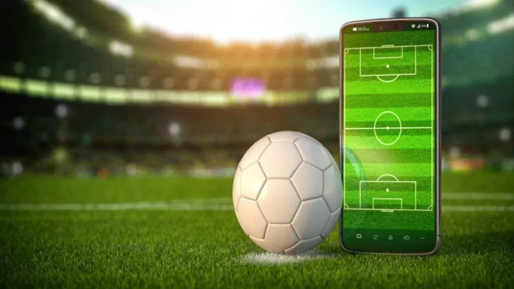Imagem de bola de futebol branca ao lado de smartphone lado a lado em gramado de estádio de futebol. 