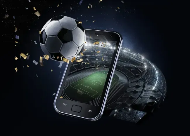 Imagem composta de fundo negro, estádio de futebol em tons escuros, celular mostrando o campo do estádio, uma bola de futebol no topo da composição