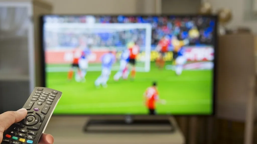 Foto de mão apontando controle remoto para TV ligada em jogo de futebol, representando alguém que aposta em casas de apostas que aceitam boleto bancário. 