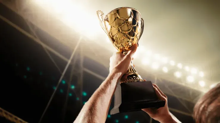 Mãos masculinas levantam troféu dourado, com luzes de arquibancada de estádio de fundo