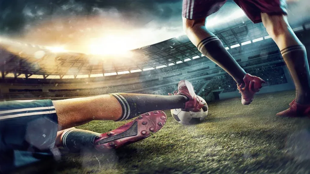 Imagem gráfica de dois jogadores do mesmo time, apenas com as pernas visíveis, indo ao encontro de bola em estádio de futebol. 