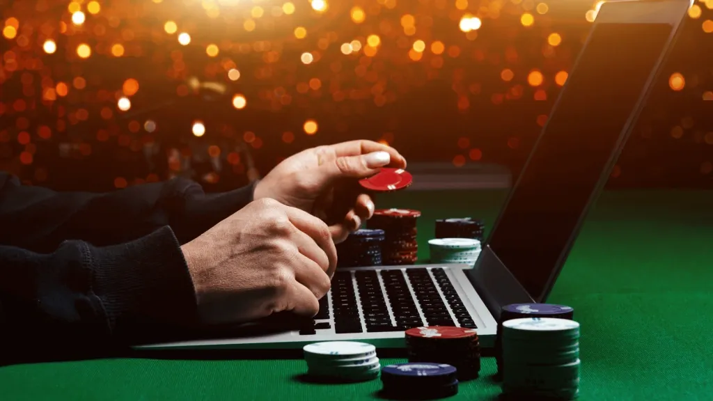 Pessoa segurando fichas de cassino em frente a notebook, com pilhas de fichas ao redor, contra fundo embaçado. 