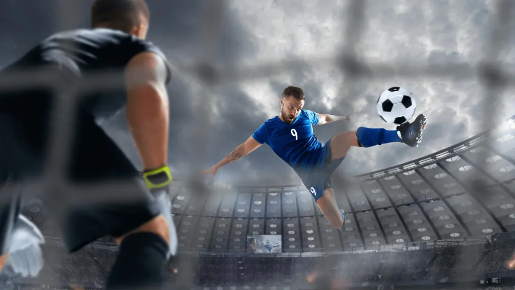 Lance de jogador ao ar, com equipagem azul, fazendo um voleio, enquanto o goleiro, de costas e equipagem preta, se prepara para tentar agarrar a bola. 