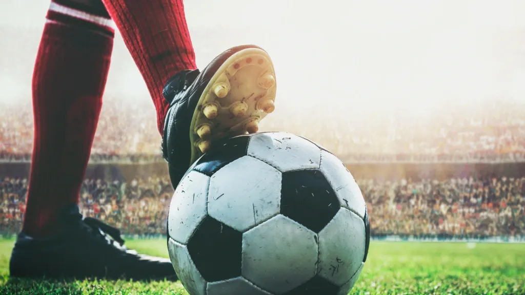 Jogador pisando em bola de futebol, apenas as pernas à vista. Ao fundo e fora de foco, estádio de futebol lotado. 