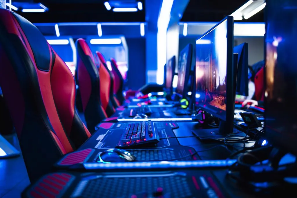 Foto de quarto iluminado com luzes neon, com setups de gamers lado a lado. O setup é composto por cadeiras com detalhes em vermelho, mouses e teclados iluminados, representando os melhores sites de apostas para e-Sports.