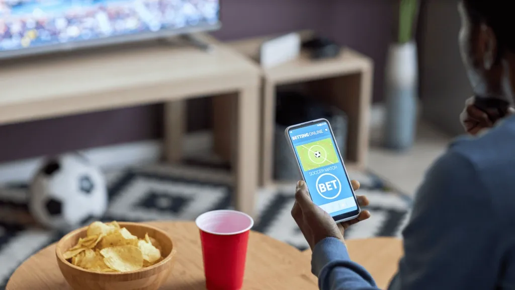 Pessoa segurando smartphone com casa de apostas na tela e em frente a uma TV, apenas metade do rosto, braço e ombro à vista. Em uma mesa redonda, à esquerda, tigela com batatas chips e um copo vermelho de bebida.