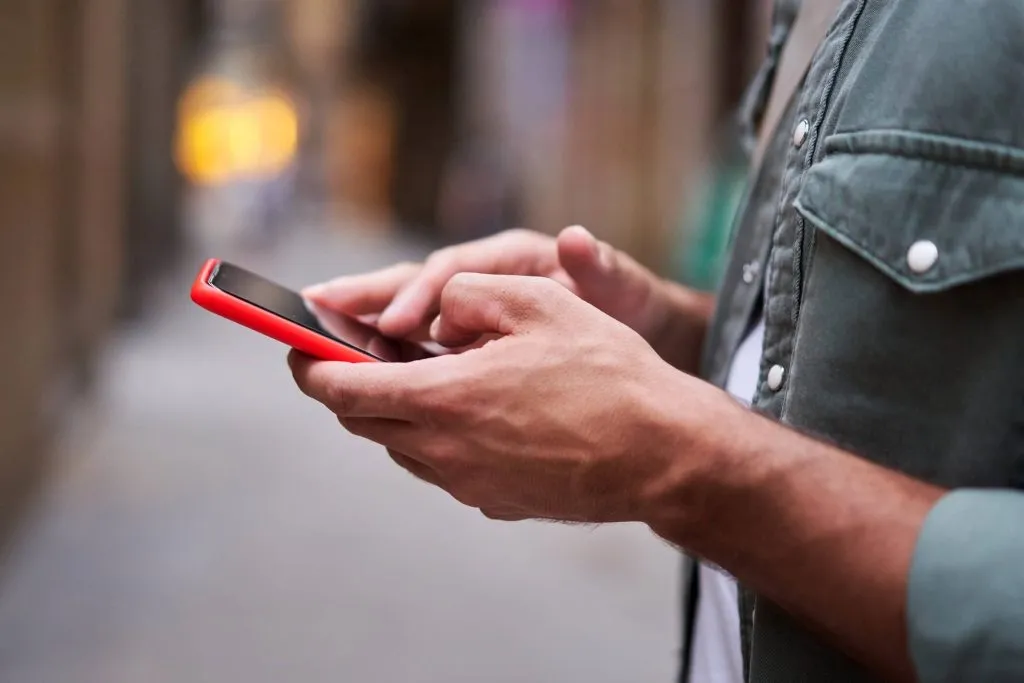 Pessoa manuseando smartphone com capa vermelha na rua com fundo embaçado. 
