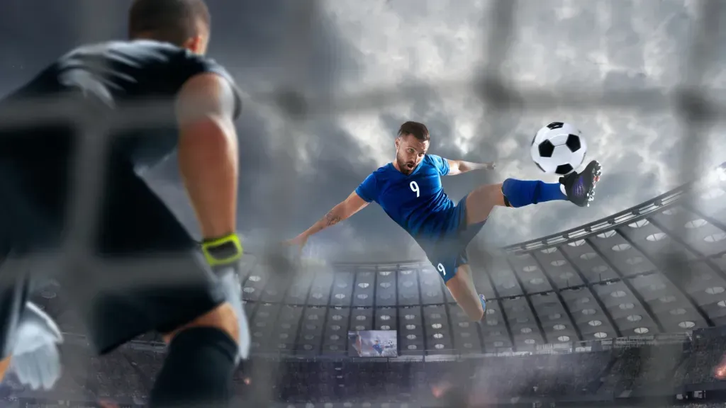 Jogador de futebol, equipado de azul, remata à baliza, com goleiro equipado de preto.