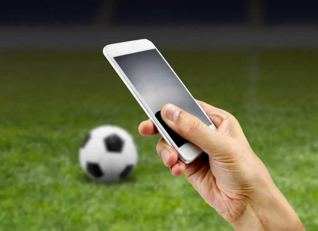 Mão segurando smartphone. Ao fundo, bola de futebol em branco e preto posando no gramado. 