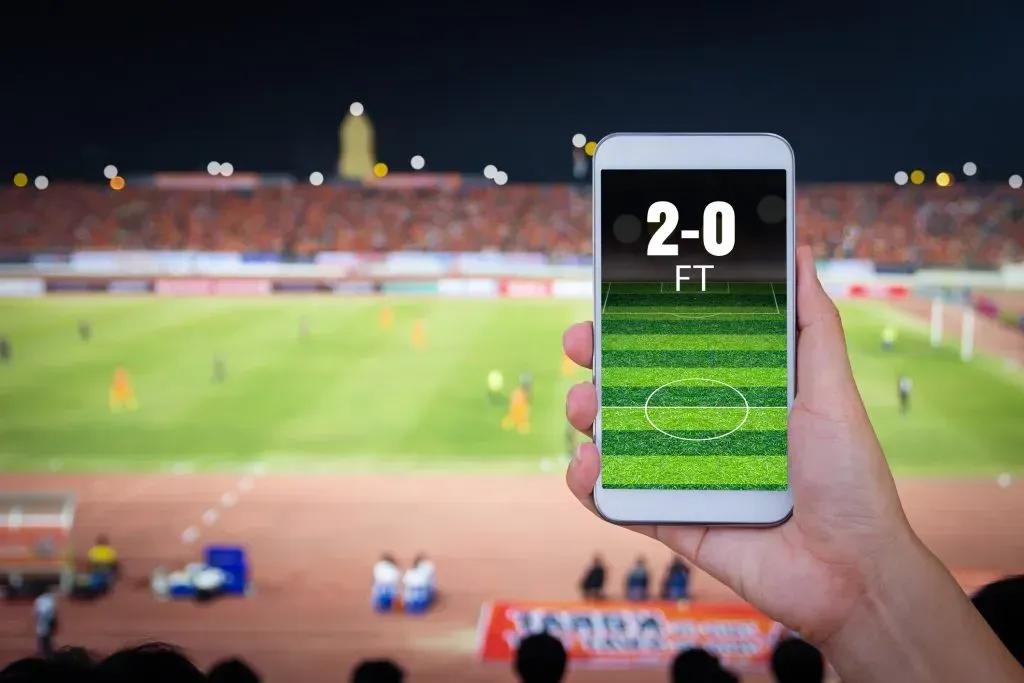 Uma mão segura um celular onde na tela se vê o placar 2-0 FT e um campo relvado, e de fundo uma arquibancada e partida de futebol a decorrer no campo do estádio.