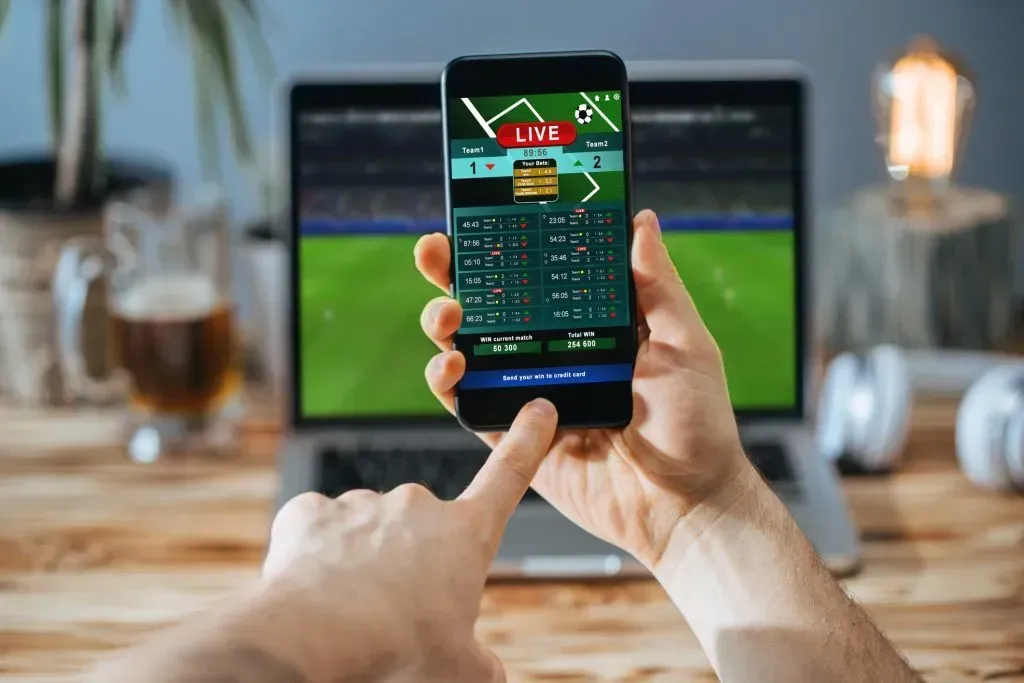 Umas mãos masculinas efetuam aposta ao vivo no celular, e no laptop de fundo passa uma partida de futebol.